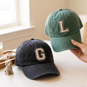 Op de afbeelding: Twee baseballpetten, één marineblauw en één groen, worden getoond. De marineblauwe pet heeft een wit omlijnde letter "G", terwijl de groene pet een wit omlijnde letter "L" heeft. Beide petten hebben een klassieke, gebogen klep.