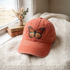 May include: A burnt orange baseball cap featuring an embroidered monarch butterfly design. The butterfly has orange, black, and white details. The cap is made of a soft material and has a curved brim.