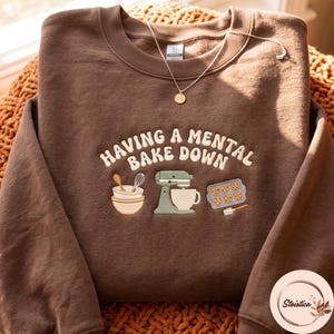 May include: A brown crewneck sweatshirt featuring embroidered text that reads "HAVING A MENTAL BAKE DOWN" in a cream-colored font. Below the text are embroidered illustrations of stacked mixing bowls, a stand mixer, and a cookie sheet.