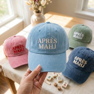 May include: A collection of baseball caps in various colors: pink, light blue, green, and navy. Each cap features the embroidered text "APR&Egrave;S MAHJ" in white. The caps are displayed on a table with mahjong tiles and dice.