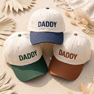 May include: Three baseball caps with the word "DADDY" embroidered on the front. The caps are cream-coloured with different coloured brims: navy blue, dark green, and brown. Each cap has a matching button on top.
