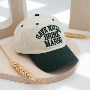 May include: Cream-colored baseball cap with a dark green brim and top button. The cap features the embroidered text "SAVE WATER DRINK MARGS" in dark green. A casual accessory for outdoor activities.