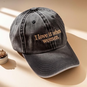 May include: A dark gray baseball cap with the embroidered text "I love it when women." The cap has a curved brim and a button on top. The hat is made of a faded, distressed fabric.