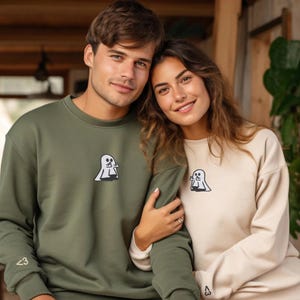 Pu&ograve; includere: Una felpa girocollo verde oliva e una felpa girocollo beige, entrambe caratterizzate da un piccolo ricamo sul petto raffigurante un fantasma bianco che fa un gesto con la mano e un piccolo simbolo "<3" ricamato sul polsino della manica.