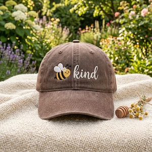 Op de afbeelding: Bruine baseballcap met het woord "kind" in witte letters geborduurd. Een cartoonbij met gele en zwarte strepen en witte vleugels is naast het woord geborduurd. De cap ligt op een beige ondergrond.