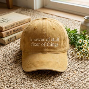 May include: Mustard-colored baseball cap with the embroidered text "knower of stuff fixer of things." The cap is made of a soft fabric and has a curved brim. The cap is sitting on a woven rug.