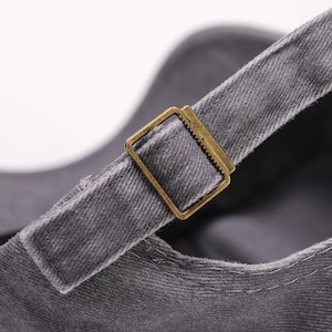 May include: Close-up of a gray baseball cap with a brass-colored metal buckle on the adjustable strap. The cap is made of a textured fabric, and the strap is sewn to the cap.