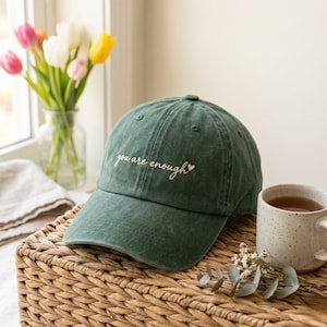 May include: A forest green baseball cap featuring the embroidered white text you are enough with a small heart symbol. The cap has a structured crown, a curved brim, and stitched eyelets, presenting a clean and simple design for daily wear.