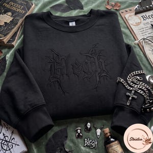 May include: A black crewneck sweatshirt featuring tonal black embroidery of the word Blegh in a stylized, jagged, gothic-inspired font across the chest. The garment has long sleeves with ribbed cuffs and a classic round neckline design.