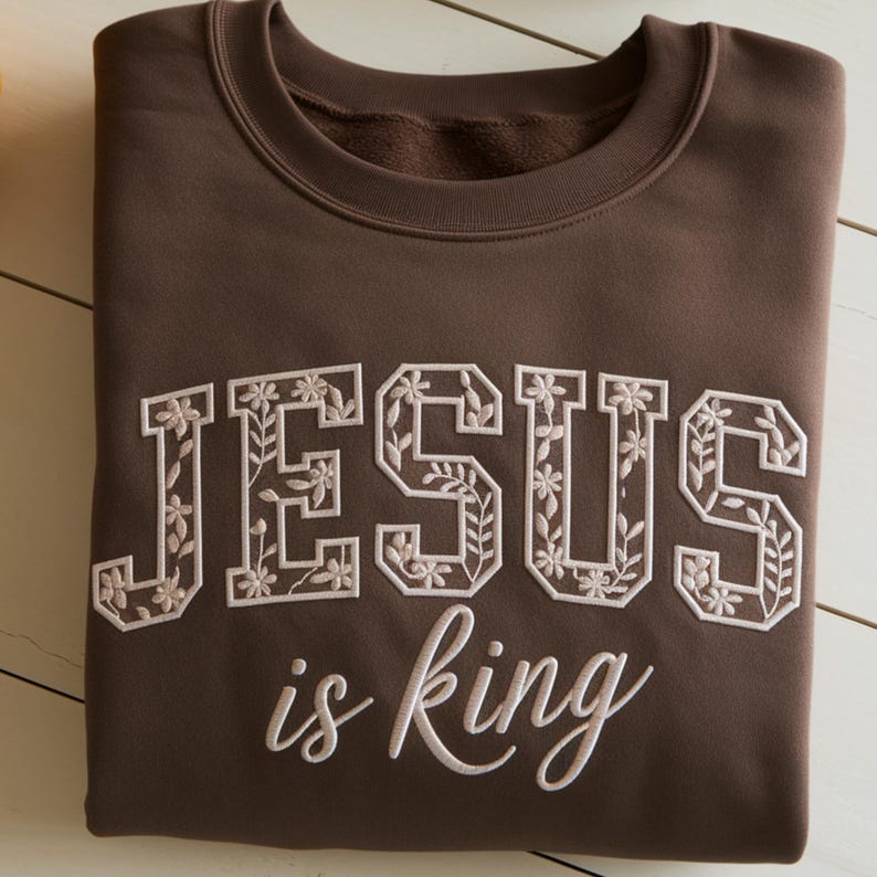 May include: A dark brown crewneck sweatshirt featuring the text JESUS in large block letters filled with a white floral and vine embroidery pattern, positioned above the words is king embroidered in a white cursive script font.