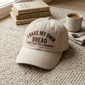 May include: A tan baseball cap with the embroidered text "I BAKE MY OWN BREAD & DON'T TRUST THE GOVERNMENT." The hat is on a textured rug, with books and a mug of coffee in the background.