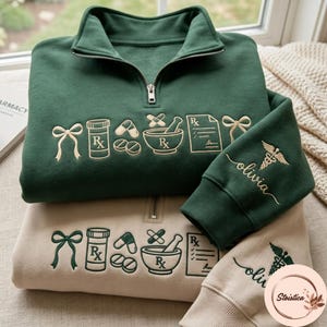 May include: A dark green and a beige quarter-zip sweatshirt, each featuring embroidered pharmacy-themed icons including a bow, pill bottle, capsules, mortar and pestle, and prescription. The sleeve displays the name Olivia and a caduceus symbol.