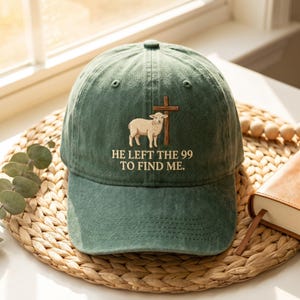 Puede incluir: Una gorra de béisbol verde bosque con un bordado que muestra un cordero blanco frente a una cruz de madera marrón. Debajo del gráfico, el texto HE LEFT THE 99 TO FIND ME aparece en letras mayúsculas de color claro.