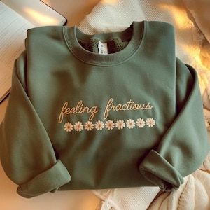 May include: Olive green sweatshirt with the embroidered phrase "feeling fractious" in a cursive font, along with a row of daisy flowers. The sweatshirt is folded, with the sleeves cuffed. The background is a soft, neutral color.