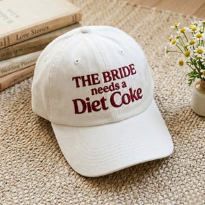 Könnte beinhalten: Weißes Baseball-Cap mit burgunderroter Stickerei "THE BRIDE needs a Diet Coke". Die Kappe liegt auf einer gewebten Oberfläche, mit Büchern und Blumen im Hintergrund.