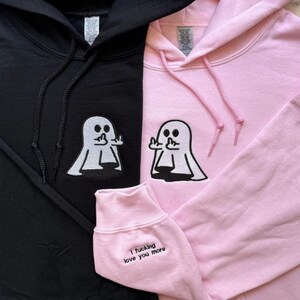 Pu&ograve; includere: Una felpa con cappuccio nera e una rosa chiaro, entrambe con un ricamo di un fantasma bianco che mostra il dito medio. La felpa rosa presenta il testo 'I fucking love you more' ricamato in nero sul polsino della manica vicino al polso.
