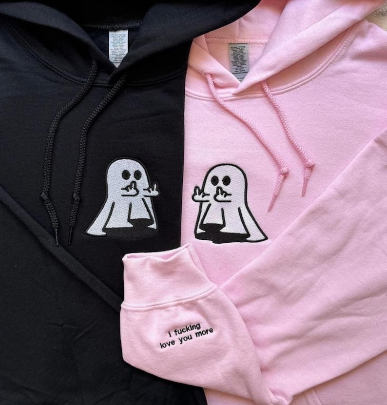 Pu&ograve; includere: Una felpa con cappuccio nera e una rosa chiaro, entrambe con un ricamo di un fantasma bianco che mostra il dito medio. La felpa rosa presenta il testo 'I fucking love you more' ricamato in nero sul polsino della manica vicino al polso.