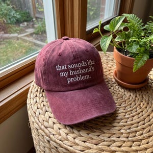 Könnte beinhalten: Eine weinrote Baseballkappe mit dem gestickten Text "that sounds like my husband's problem." Die Mütze liegt auf einer gewebten Oberfläche, mit einem Fenster und Topfpflanzen im Hintergrund.