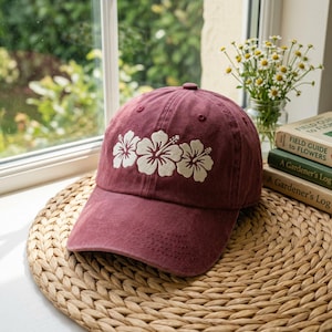 Może przedstawiać: Bordowa czapka bejsbolowa z trzema białymi kwiatami hibiskusa. Czapka wykonana jest z miękkiego materiału i ma zakrzywiony daszek. Kapelusz leży na plecionej macie w pobliżu okna.