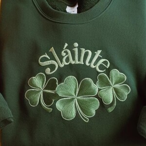 May include: Dark green sweatshirt with the embroidered word "Sl&aacute;inte" above three green shamrocks. The embroidery is in a lighter shade of green, creating a textured effect. The sweatshirt is a crew neck style.