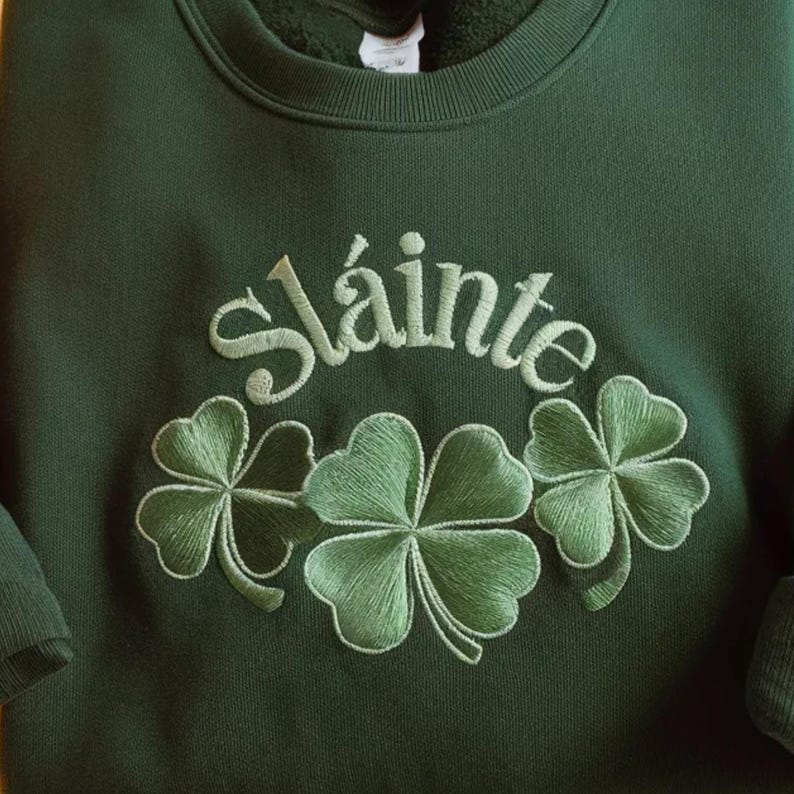 May include: Dark green sweatshirt with the embroidered word "Sl&aacute;inte" above three green shamrocks. The embroidery is in a lighter shade of green, creating a textured effect. The sweatshirt is a crew neck style.