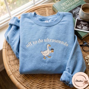 Peut inclure: Un sweat-shirt bleu clair à col rond avec une broderie représentant une oie blanche tenant une tasse de café. Le texte "off to do ultrasounds" est brodé en blanc au-dessus de l'oie dans une police minuscule et incurvée sur la poitrine.