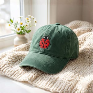 May include: A forest green baseball cap featuring an embroidered red ladybug with black spots and a black head. The cap has a curved brim, a structured front panel, and stitched eyelets on the crown, showcasing a classic six-panel design.