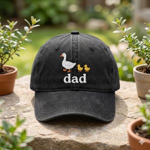 May include: A black baseball cap featuring an embroidered graphic of one white adult goose followed by two small yellow ducklings. Below the birds, the word "dad" is embroidered in white lowercase letters. The cap has a curved brim and eyelets.