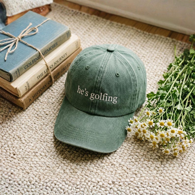 May include: A green baseball cap with the embroidered text "he's golfing" in white. The hat is on a beige rug next to a stack of books and a bouquet of white flowers.