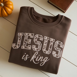 May include: A dark brown crewneck sweatshirt featuring the embroidered text "JESUS" in large block letters filled with a white floral pattern, followed by the word "is king" in a smaller, white cursive script font centered below.