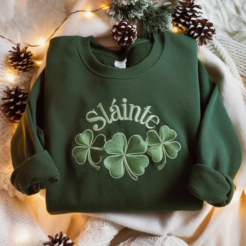 May include: A dark green sweatshirt with the embroidered word "Sl&aacute;inte" above three embroidered shamrocks. The sweatshirt has a crew neck and cuffed sleeves. The background includes pine cones and greenery.