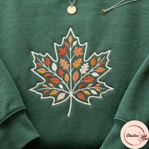 May include: A dark green sweatshirt featuring a large embroidered maple leaf design. The leaf is outlined in white and filled with smaller, multi-colored leaves in shades of orange, yellow, and cream. A gold necklace is visible at the top.