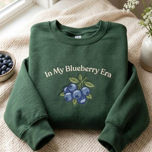 May include: A dark forest green crewneck sweatshirt featuring an embroidered graphic of a cluster of blueberries with green leaves. The text "In My Blueberry Era" is embroidered in a cream-colored font above the blueberry illustration.