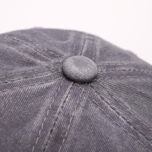May include: Close-up of a gray newsboy cap with a rounded top and a small, matching button. The cap is made of a textured fabric with visible stitching details. The hat is a neutral color, suitable for various outfits.