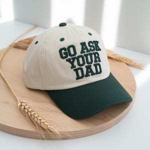 Peut inclure: Casquette de baseball beige et vert fonc&eacute; avec le texte brod&eacute; "GO ASK YOUR DAD". La casquette a une visi&egrave;re vert fonc&eacute; et un bouton sur le dessus. Le chapeau est pos&eacute; sur un plateau en bois.