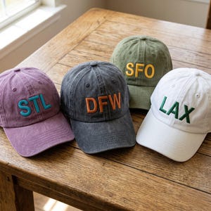 May include: Four baseball caps in various colors, each embroidered with a three-letter airport code: STL, DFW, SFO, and LAX. The caps are in shades of purple, charcoal, olive green, and white, with contrasting embroidery.
