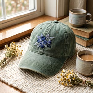 May include: A green baseball cap with an embroidered design of blueberries and leaves. The cap is made of a soft, worn fabric and has a curved brim. The embroidery features shades of blue and gray.