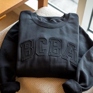 Peut inclure: Un sweat-shirt noir avec les lettres "BCBA" en relief et texturées sur la poitrine. Le sweat-shirt est plié avec les manches rentrées. Le tissu semble doux et confortable.