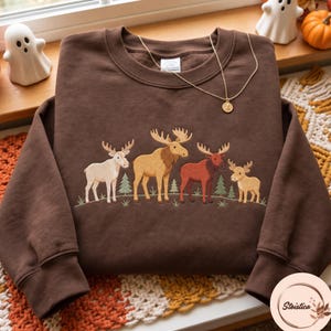 May include: A dark brown crewneck sweatshirt featuring an embroidered design of four moose of varying sizes and colours standing among small green pine trees. A thin gold-toned chain necklace with a small round pendant rests on the neckline.