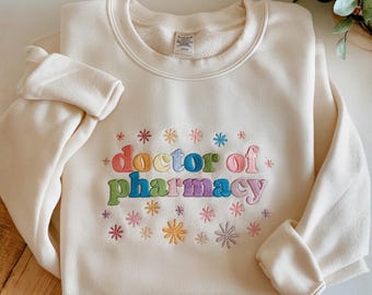 Könnte beinhalten: Ein cremefarbenes Sweatshirt mit Rundhalsausschnitt und dem gestickten Text "doctor of pharmacy" in bunten Buchstaben. Das Design ist von kleinen, farbenfrohen, stern- oder blumenartigen Stickereien in Rosa-, Blau- und Gelbtönen umgeben.