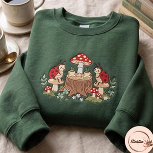 Puede incluir: Una sudadera de cuello redondo en color verde oscuro con un bordado que muestra a dos mariquitas sentadas sobre setas en una mesa de tronco de árbol. El diseño incluye una seta grande roja con puntos blancos, dos tazas humeantes y flores.