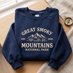 May include: A navy blue crewneck sweatshirt featuring a cream-coloured graphic of the Great Smoky Mountains with the text "GREAT SMOKY MOUNTAINS NATIONAL PARK EST. 1940" and small green pine trees. The design is centred on the chest of the garment.