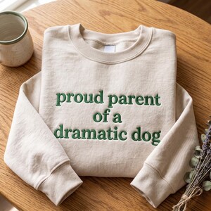 May include: A beige crewneck sweatshirt featuring dark green embroidered text on the chest that reads, "proud parent of a dramatic dog." The garment has long sleeves, a ribbed collar, and ribbed cuffs, showcasing a clean and simple design.