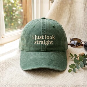 May include: A forest green baseball cap with a curved brim and an adjustable strap at the back. The front of the cap features the embroidered text "i just look straight" in a light cream colour, centred in a simple, clean, sans-serif font.