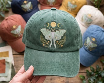 May include: A forest green baseball cap featuring an embroidered design of a pale green moth centred below a yellow sun and two crescent moons. The moth is flanked by green fern fronds and small yellow flowers, with tiny stars scattered above.