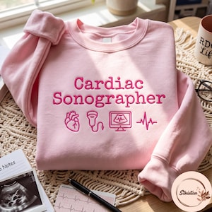 Puede incluir: Una sudadera de cuello redondo rosa claro con bordado rosa oscuro que dice Cardiac Sonographer. Debajo del texto hay iconos bordados de un corazón humano, un transductor de ultrasonido, un monitor con una onda y una línea de ritmo cardíaco EKG.