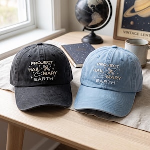 May include: Two baseball caps, one in charcoal black and one in light blue, each featuring white embroidered text that reads PROJECT HAIL MARY EARTH, accompanied by a small satellite graphic and star symbols centered on the front panels.