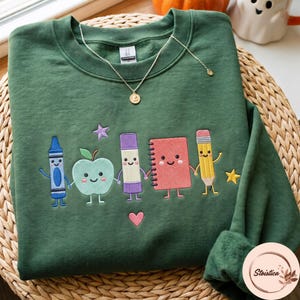 Könnte beinhalten: Ein waldgrüner Rundhals-Pullover mit einer Stickerei, die einen blauen Buntstift, einen hellgrünen Apfel, einen violetten Klebestift, ein rotes Notizbuch und einen gelben Bleistift zeigt, die sich an den Händen halten, ergänzt durch ein kleines rosa Herz.