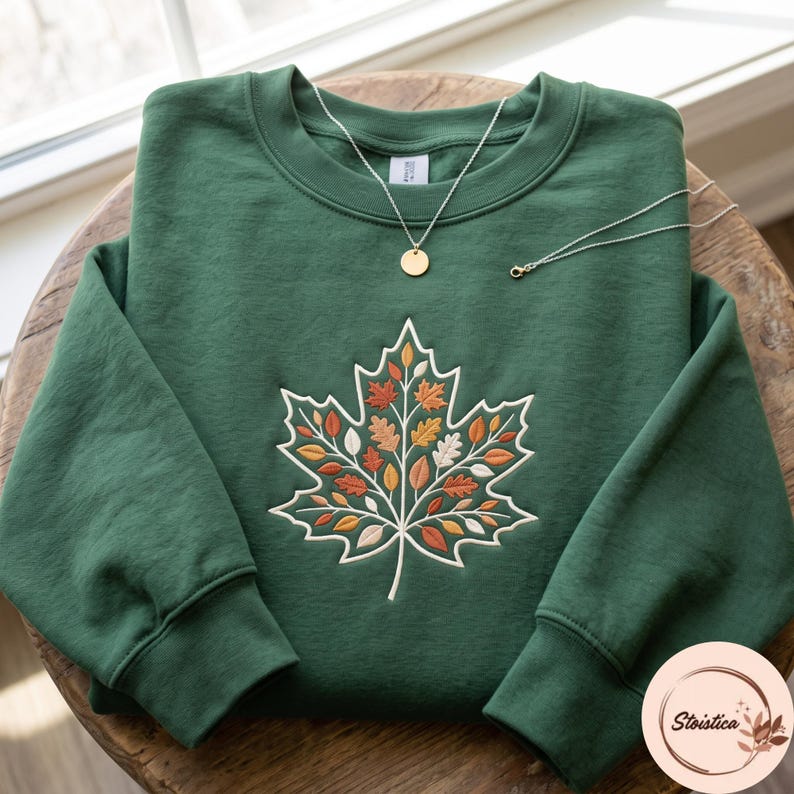 May include: A forest green crewneck sweatshirt featuring a large embroidered maple leaf design on the chest. The leaf is filled with smaller, multi-colored autumn leaves in shades of orange, yellow, and cream. A gold-tone necklace rests on top.