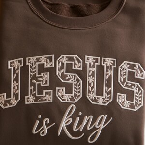 May include: A dark brown crewneck sweatshirt featuring the word JESUS in large block letters filled with an embroidered white floral and vine pattern, positioned above the words is king in a white cursive script font.